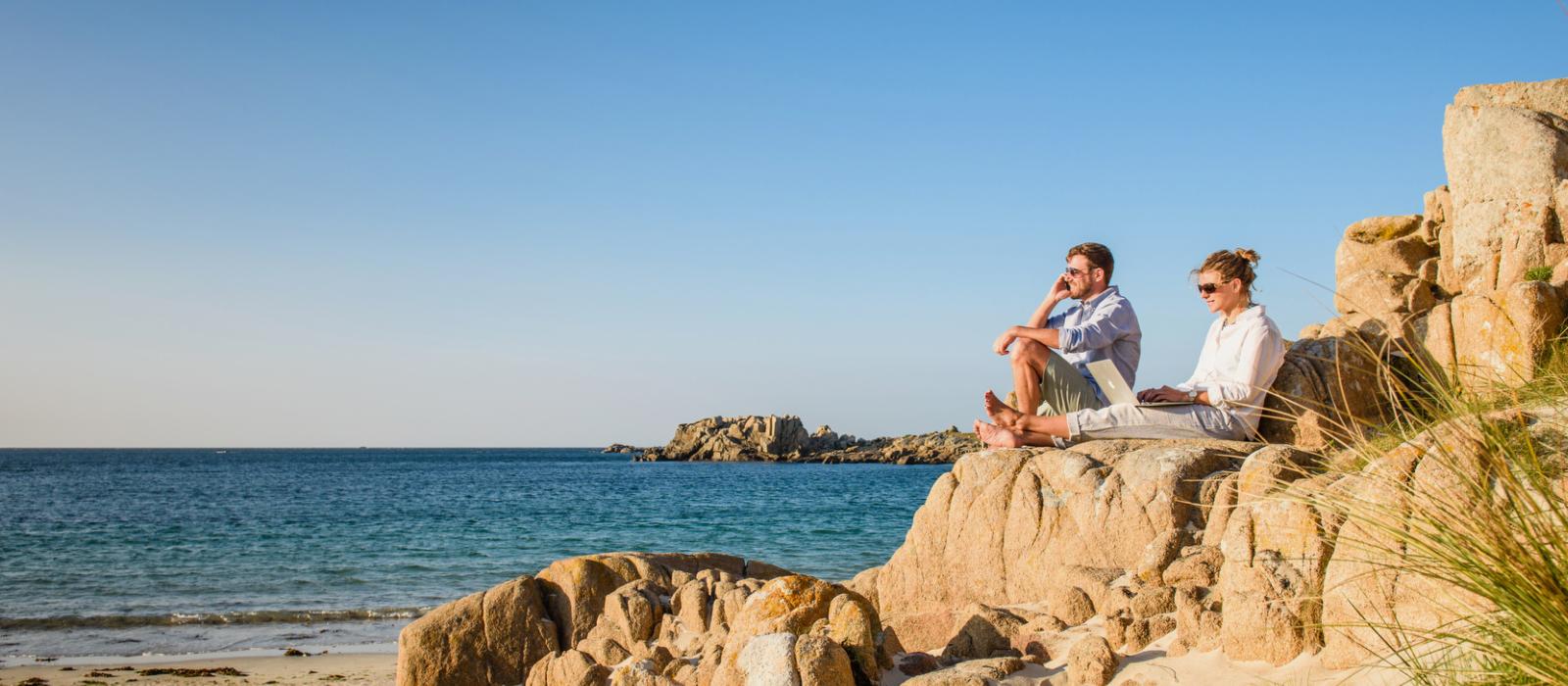 Two people sat on a rocky ledge on the beach. the man is on the phone whilst the woman is on her laptop