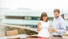 Two people stood on a balcony gazing at an ipad and smiling